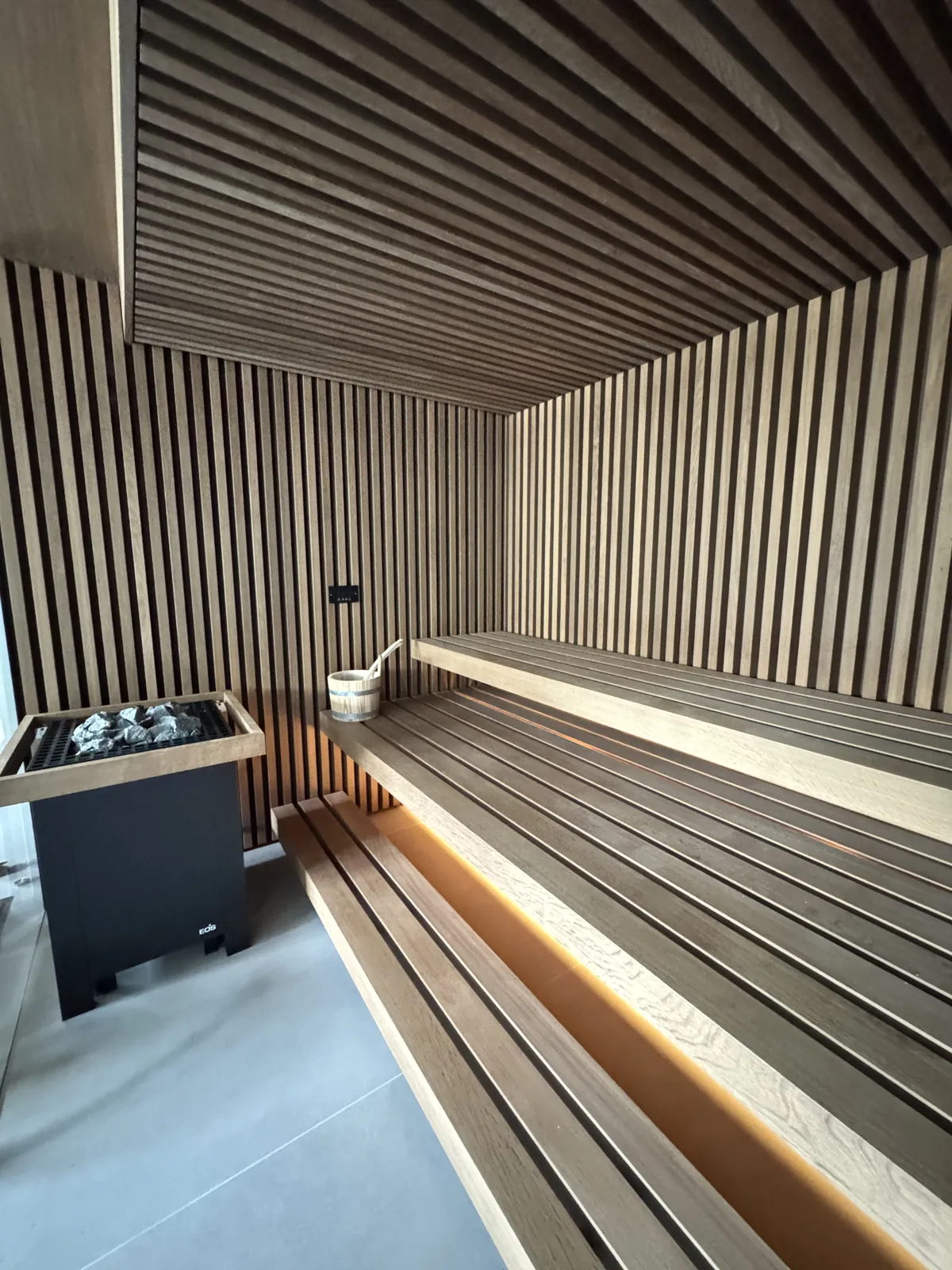 Designsauna mit dunklen Holzlamellen an Wänden und Decke, beleuchteten Sitzbänken und EOS-Saunaofen